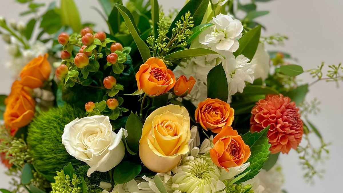 Fall & Thanksgiving flowers online. A vibrant autumnal floral arrangement featuring orange and yellow roses, white roses, a coral dahlia, green foliage, and small berry clusters at FIORI Florist in Oakville